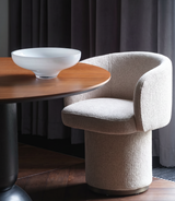Beige chair next to a wooden table with a bowl on it, against a dark curtain background.